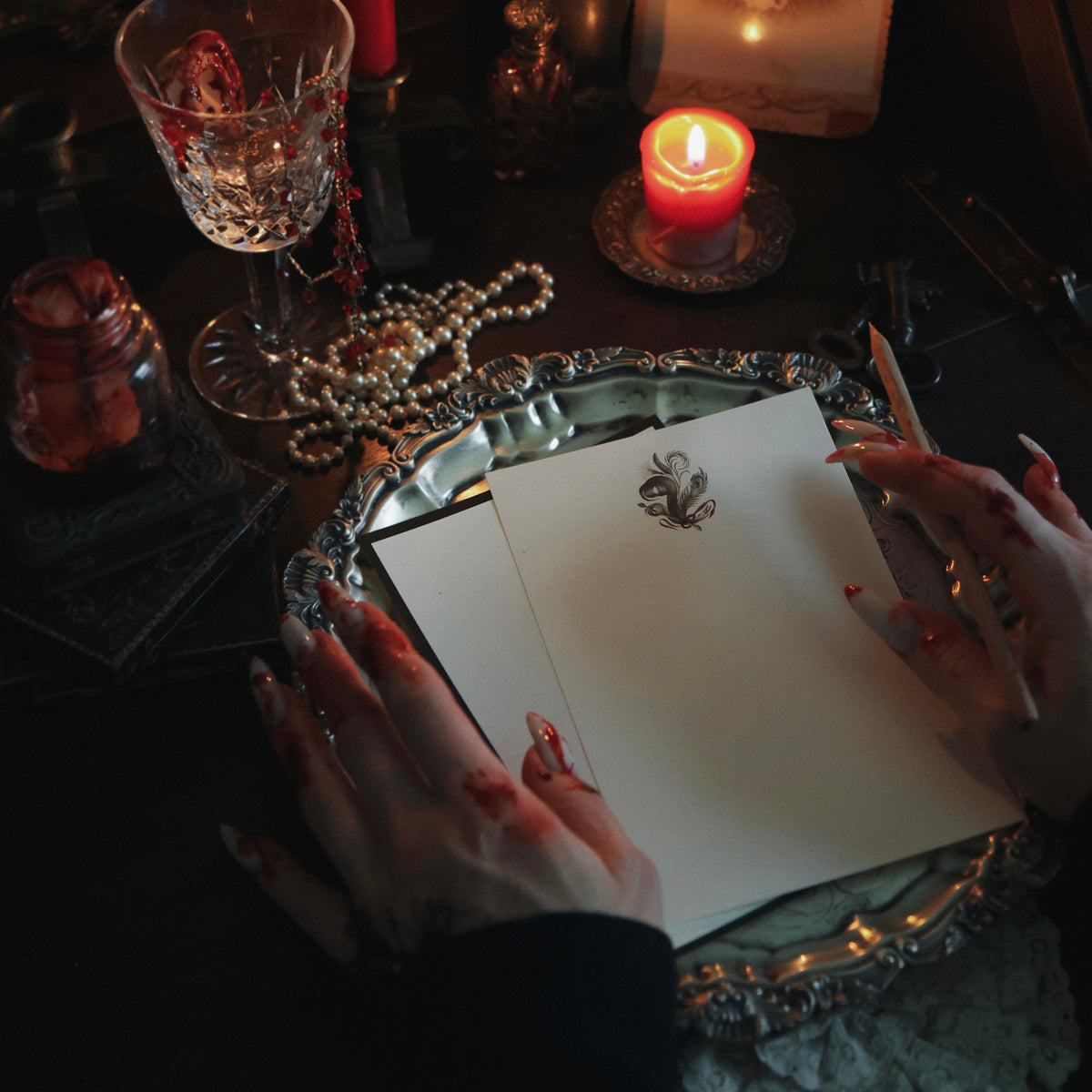 A dark, moody tabletop scene with two bloody hands on a gothic letter writing set with candles, jewelry, and horror-themed props.