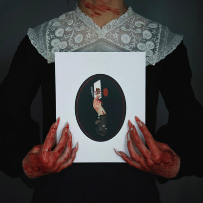 Bloody woman in a Victorian dress with long fingernails holds a gothic collage art print featuring a bloody hand with a razor, a note, and an eye.